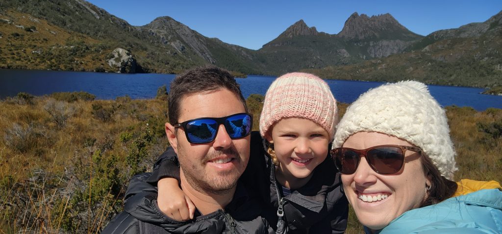 Happy family travelling Tasmania Australia, standing in front of Dove Lake, Cradle Mountain wearing cold weather clothes, in sunshine. 