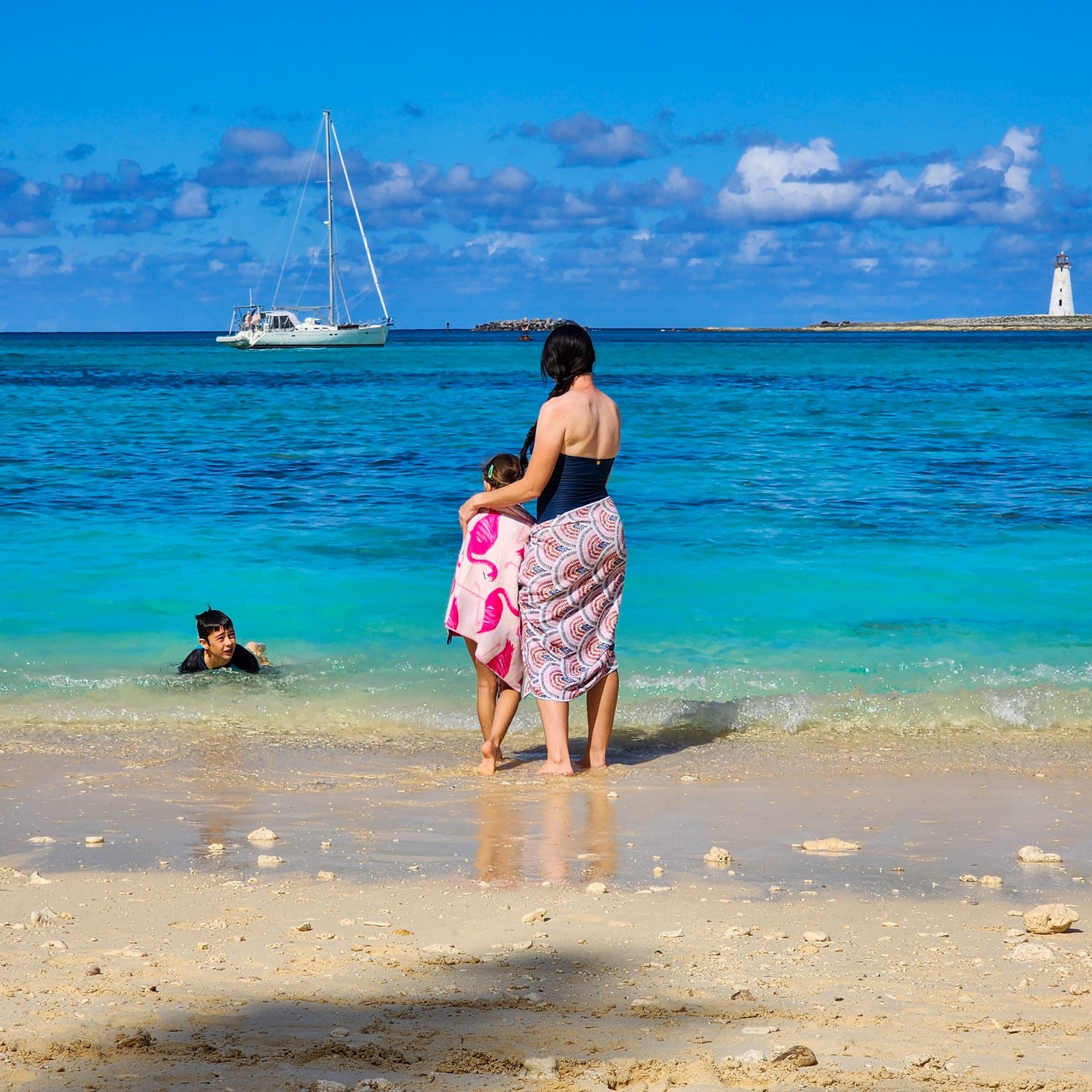 Mother and daughter standing at the beach, lookout out at the blue water with beach towels around their waists.